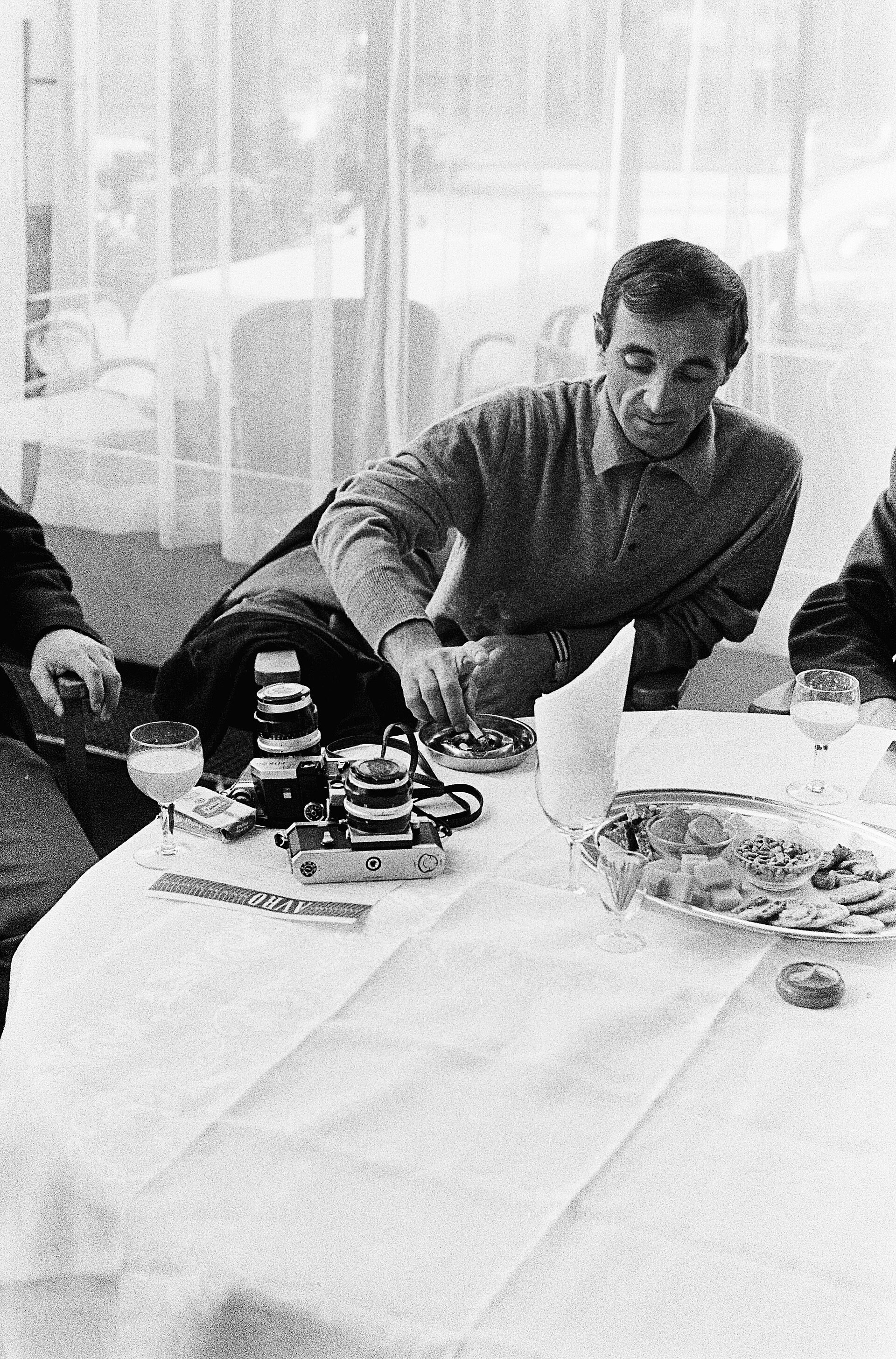 Aznavour at a hotel table in the Netherlands, cameras and wine glasses beside him